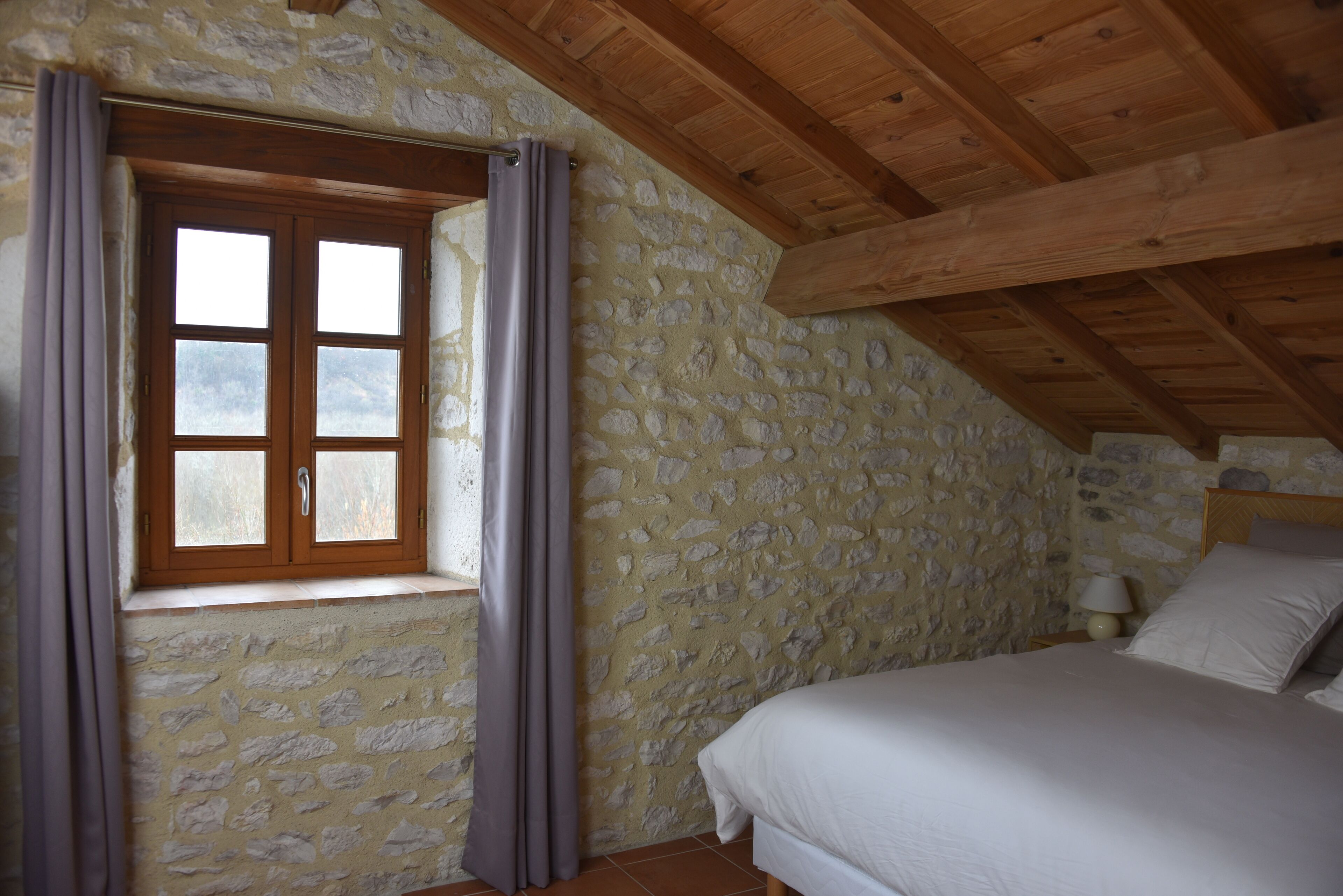Ein Schlafzimmer mit Steinsandsteinwand, Holzbalkendecke, einem Fenster mit Vorhängen, einem Bett mit weißer Bettwäsche und einer Nachttischlampe.