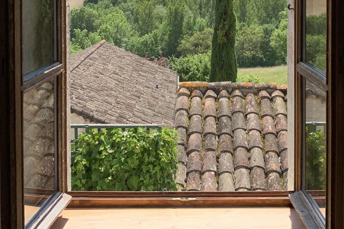 Uma janela aberta com vista para uma paisagem verde, um telhado em telhas e uma árvore alta.