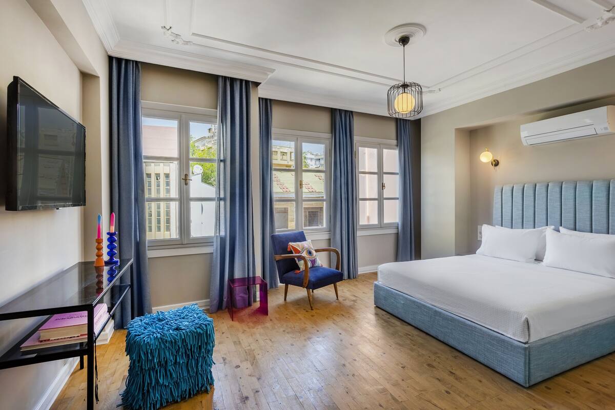 Quarto de hotel com uma cama grande, televisão, cadeira, mesa pequena e janela com cortinas.