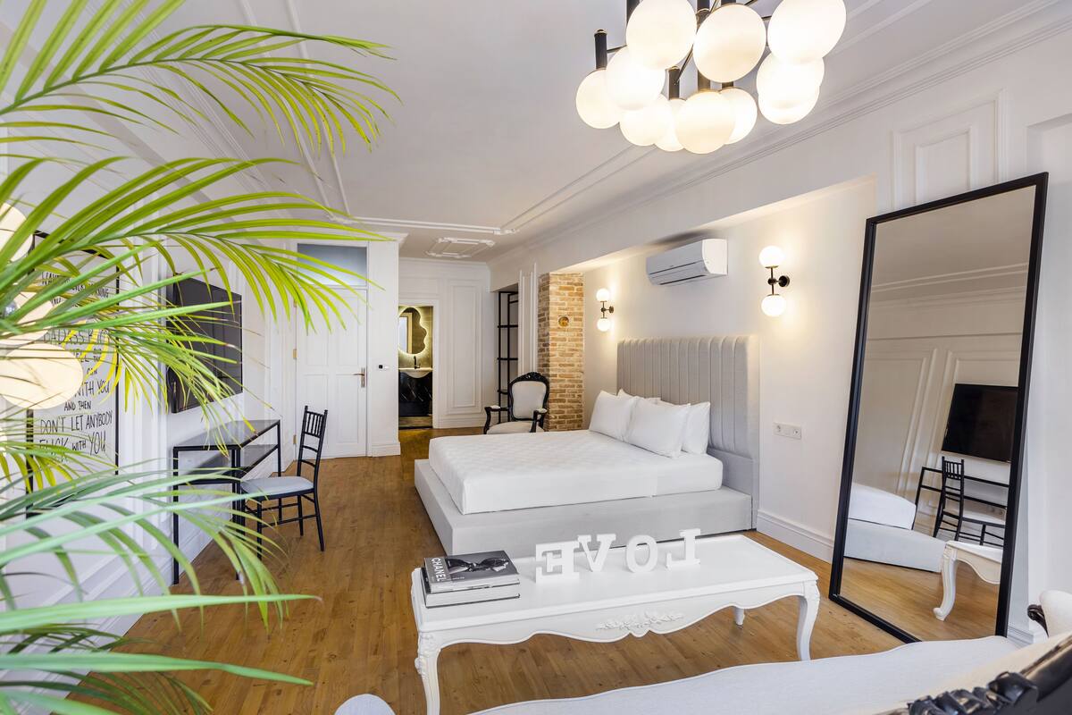 Quarto moderno com uma cama grande, um espaço de estar com mesa e um espelho amplo.
