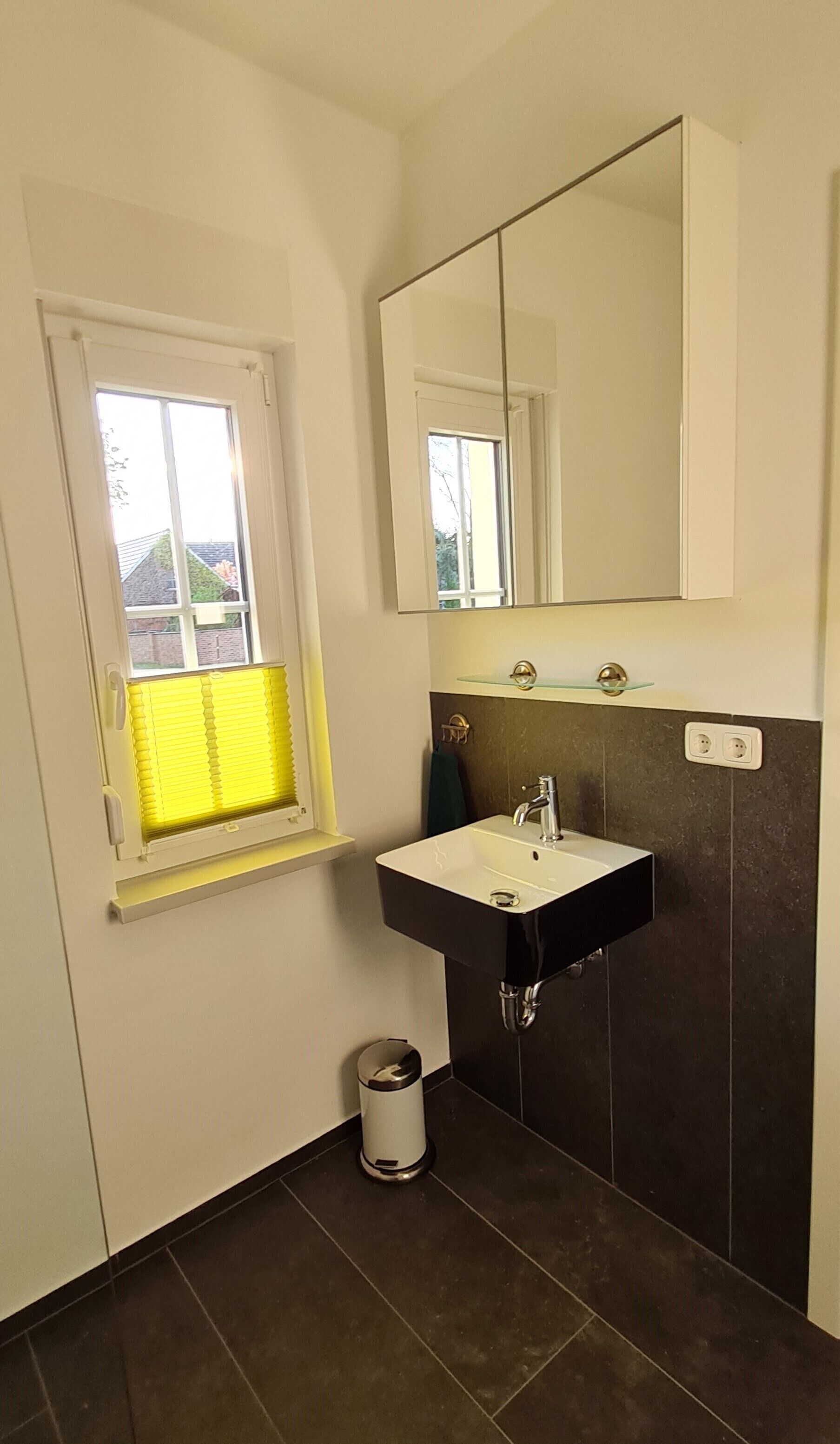 A small bathroom with a window, a sink, a mirror, and a trash bin.