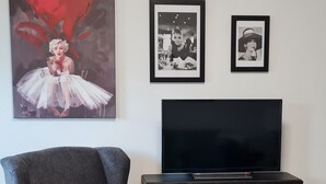 A living room with a dark grey armchair, a matching ottoman, a flat-screen TV on a wooden cabinet, and three framed pictures on the wall.