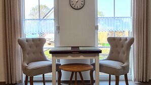A dining area with a table and two chairs, a wall clock, and large windows with curtains.