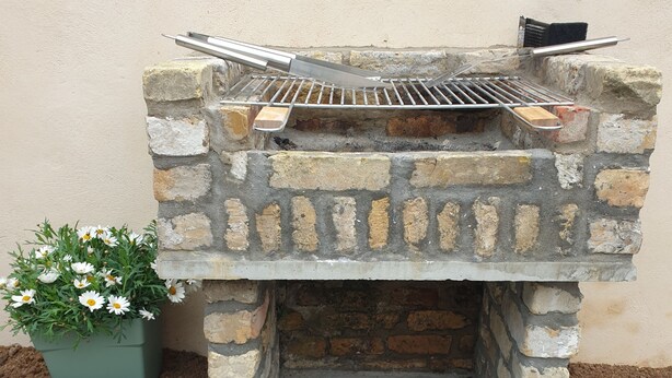A stone-built outdoor grill with a metal grate and a brick base, set against a wall with a potted plant nearby. - Cosy detached house - 4 people - 2 bedrooms Terrace (Bourbourg)