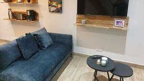 A living room with a blue sofa, a flat-screen TV mounted on a wooden shelf, a coffee maker, a toaster, and a small table with two cups and a remote control. - Cosy detached house - 4 people - 2 bedrooms Terrace (Bourbourg)