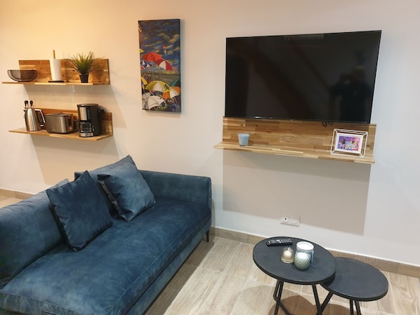 Een woonkamer met een blauwe bank, een flatscreen televisie bevestigd aan een houten plank, een koffiezetapparaat, een broodrooster en een kleine tafel met twee kopjes en een afstandsbediening.