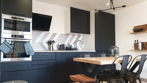 A modern kitchen with a built-in oven, microwave, and a large screen above the oven. There is a wooden dining table with chairs and a trash bin. - Cosy detached house - 4 people - 2 bedrooms Terrace (Bourbourg)