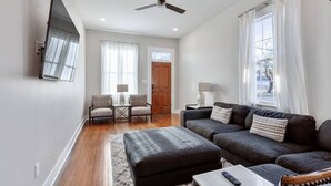 A living room with a dark sofa set, a coffee table, a TV mounted on the wall, and a ceiling fan. - Historical Charm with Modern Touch 5 Bedrooms 4 Bathrooms, Centrally Located (New Orleans)