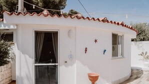 Een klein, laag gebouw met een witte gevel en terracotta dakpannen. Het heeft een groot raam met gordijnen, een kleine deur en een bloempot op de grond.
