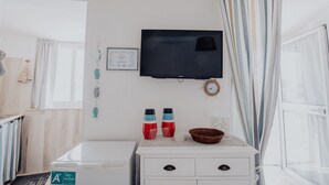 Een kamer met een wandgemonteerde flatscreen televisie, een witte dressoir, een klein tafeltje met een mand en een raam met gordijnen.