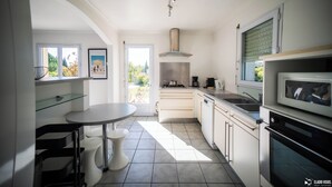 A modern kitchen with white cabinets, a stainless steel range hood, a microwave, and a dining area with a round table and chairs. - Villa (Poulx)