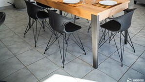 A dining area with a wooden table set for four, surrounded by black chairs. There are potted plants on either side of the table, and a door with a glass panel is visible in the background.