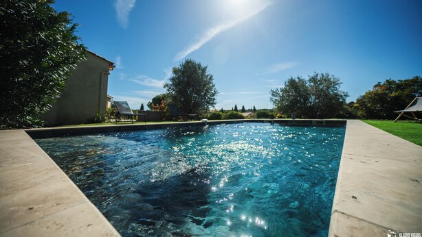 A rectangular swimming pool with clear water, surrounded by a concrete deck and a grassy area with trees and a building in the background. - Villa (Poulx)