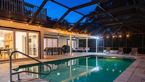 An indoor swimming pool with a glass enclosure, a covered patio area with seating, and a view of the surrounding trees.