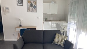 Ett modernt vardagsrum med en grå soffa, en mönstrad kudde, ett skrivbord med en stol och ett kök i bakgrunden.
