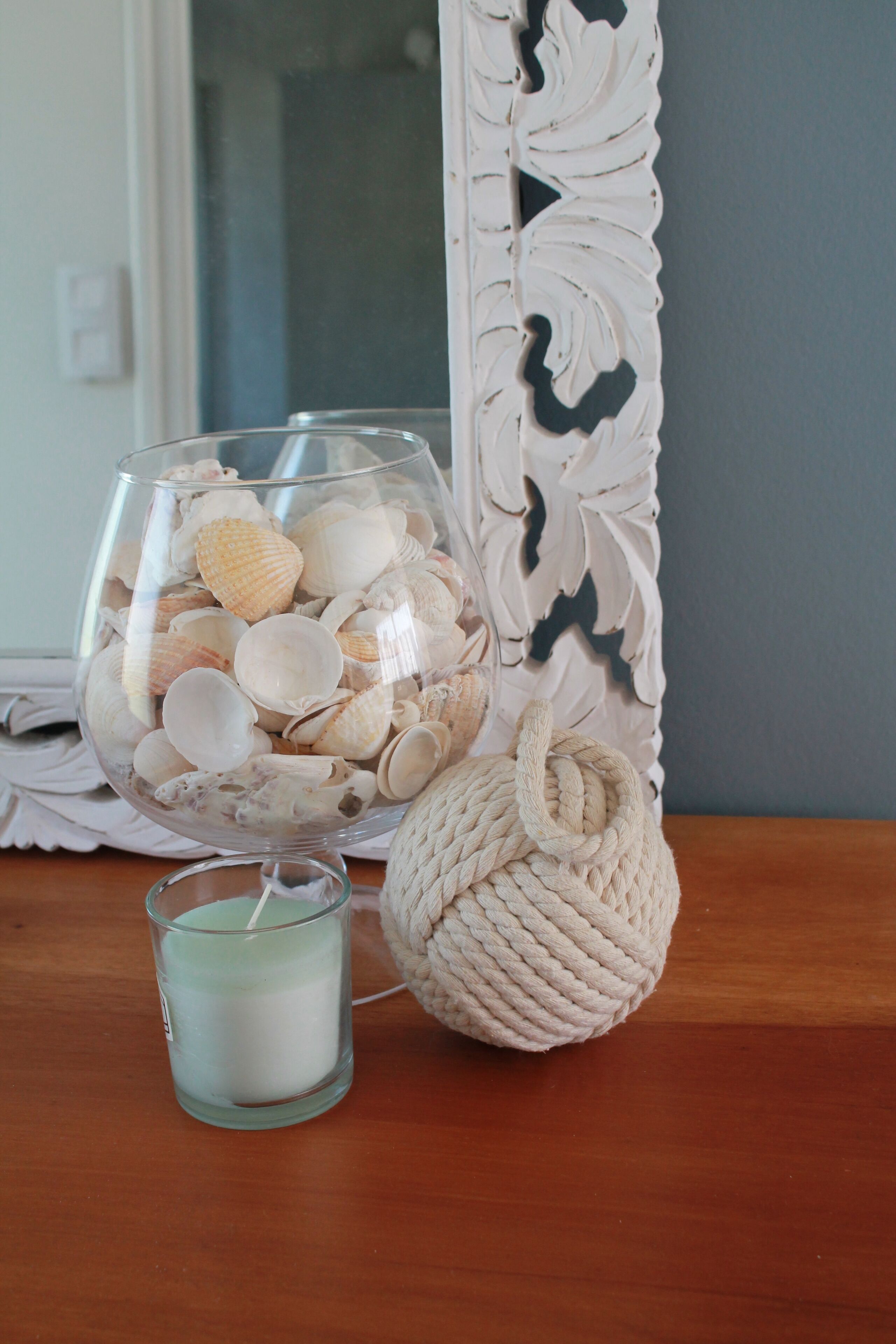 A glass bowl filled with seashells, a small candle, and a knitted ball on a wooden surface.