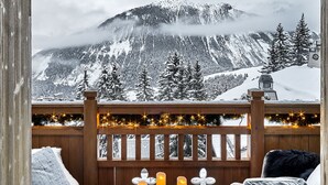 A cozy outdoor seating area with two cushioned chairs, a round table with candles and pastries, and a view of snow-covered mountains. - LYS MARTAGON (Courchevel)