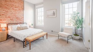 Ein Schlafzimmer mit Backsteinwand, einem Bett mit hölzernem Sitzbrett, einem weißen Stuhl und einem Blumentopf.