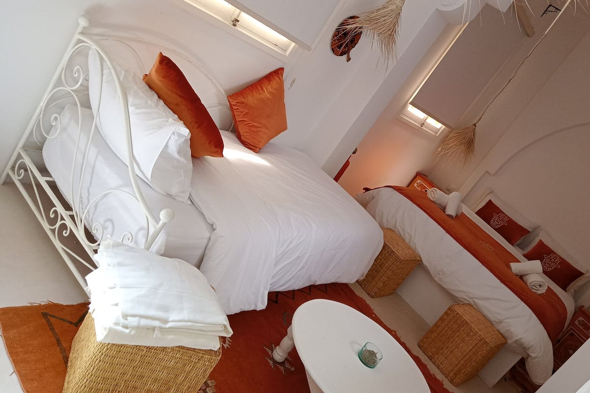 A bed with white and orange bedding, a small table with a glass, and a wicker basket.