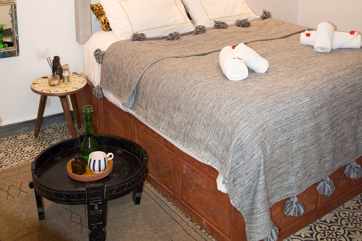 A neatly made bed with a grey blanket, two pillows, and two rolled towels. There is a small round table with a bottle and a cup beside the bed.