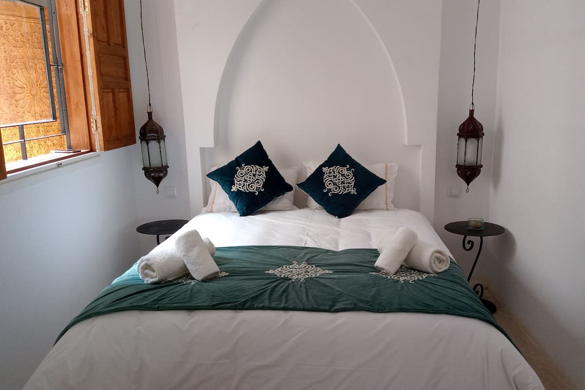A neatly made bed with two decorative pillows, a green bedspread, and two white towels.