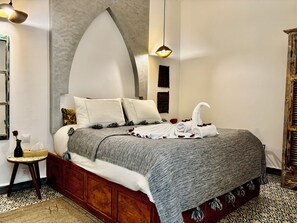 A neatly made bed with a grey and white patterned blanket, a wooden headboard, and a small bedside table with a vase and a tissue box. - Le Khan Mogador (Essaouira)