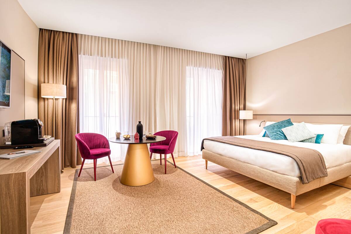 A hotel room with a bed, a desk, a chair, a table, and a large window with curtains.