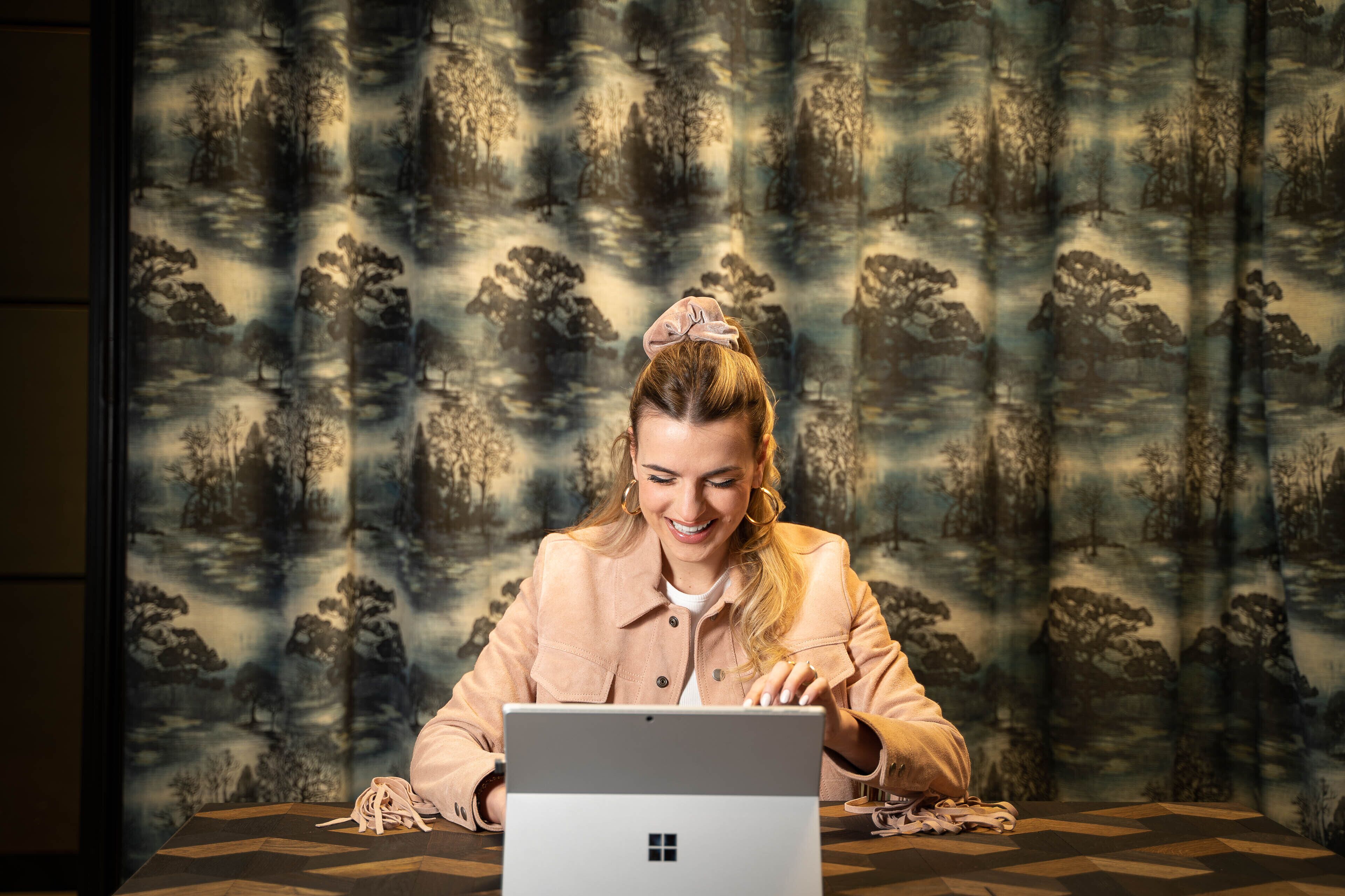 A person sitting at a table with a laptop, a patterned curtain in the background, and a geometric patterned tablecloth.