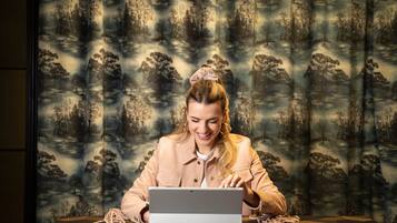 A person sitting at a table with a laptop, a patterned curtain in the background, and a geometric patterned tablecloth.