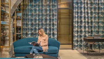 A woman sitting on a blue sofa reading a book in a room with a bookshelf, a table with a laptop, and a curtain with a tree pattern.