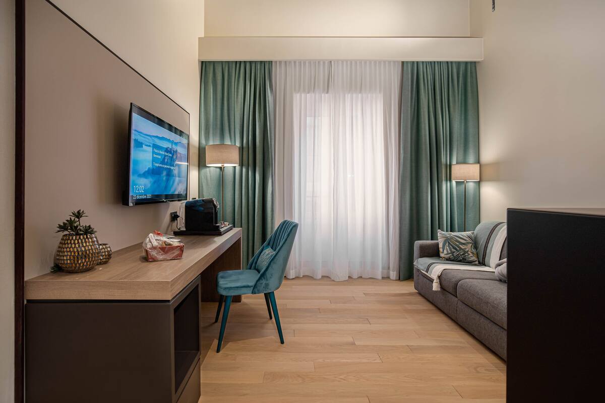 A hotel room with a flat-screen TV mounted on the wall, a desk with a laptop, a chair, a sofa, and two lamps.