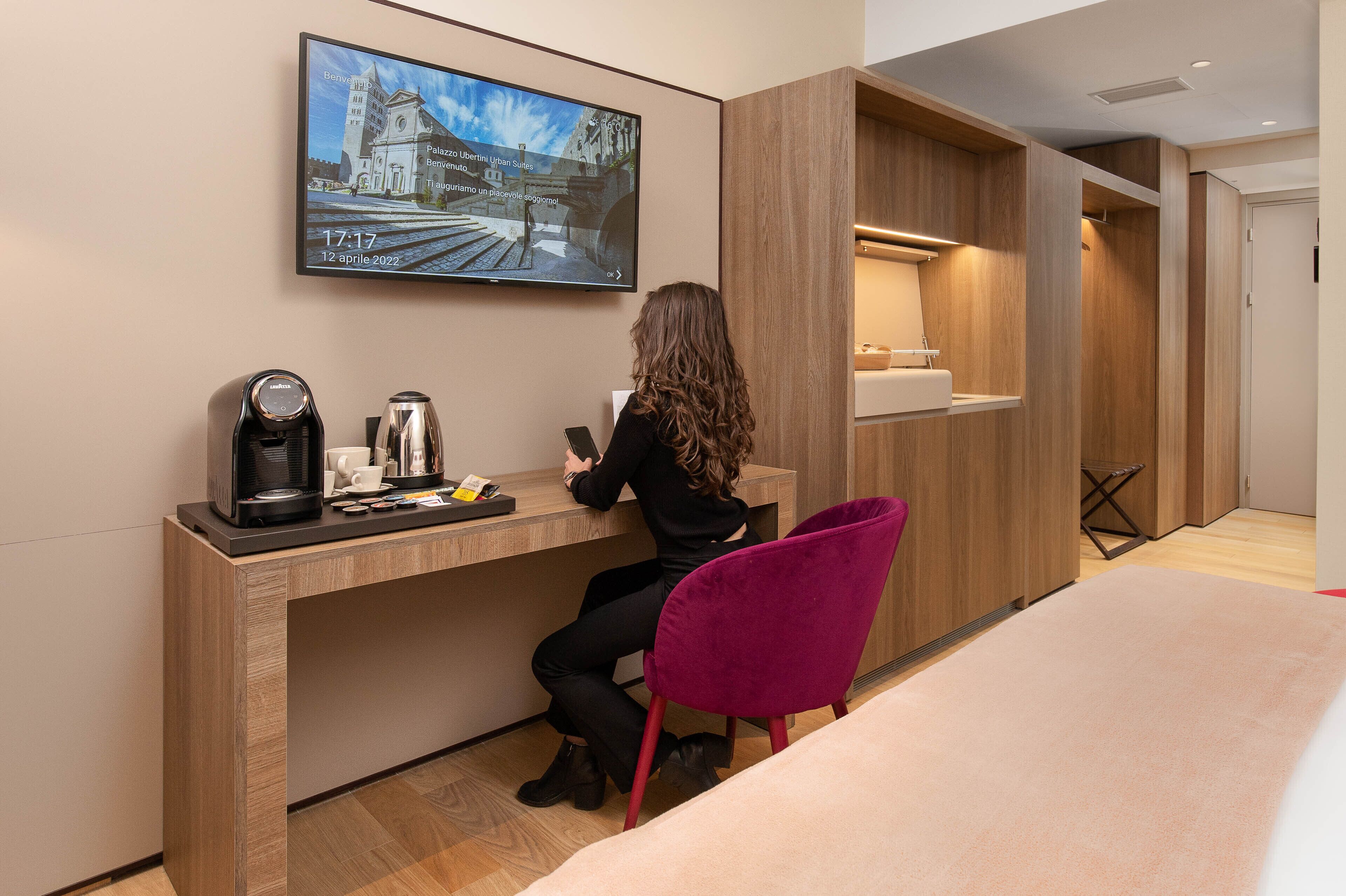 A hotel room with a wooden desk, a coffee maker, a laptop, and a television displaying a cityscape.
