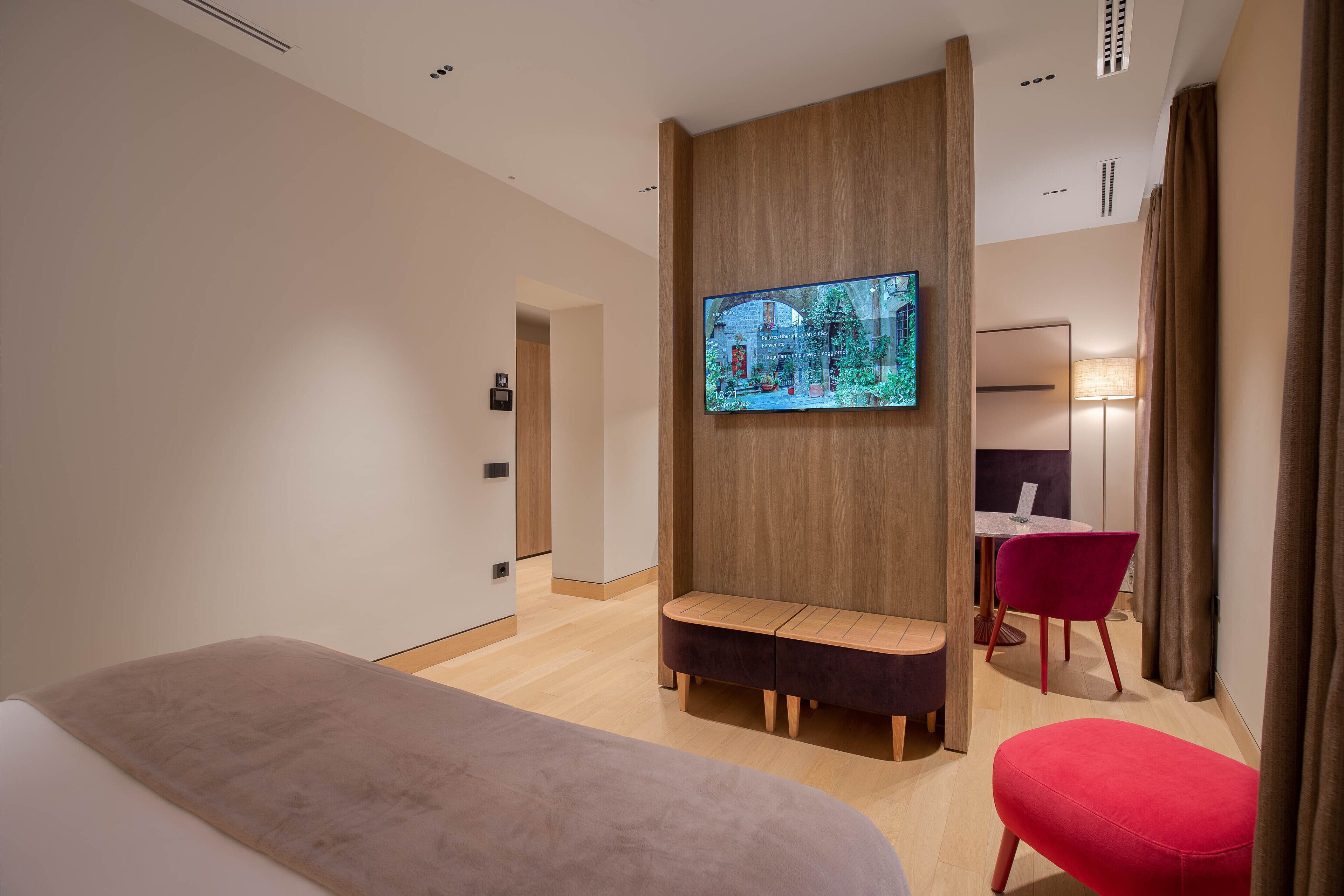 Une chambre d’hôtel moderne avec un grand lit, une cloison en bois sur laquelle est fixée une télévision, un fauteuil rouge et une petite table avec une lampe.