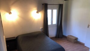 Ein Zimmer mit einem Bett, einer kleinen Bank, einem Fenster mit Vorhängen und zwei Wandleuchten.
