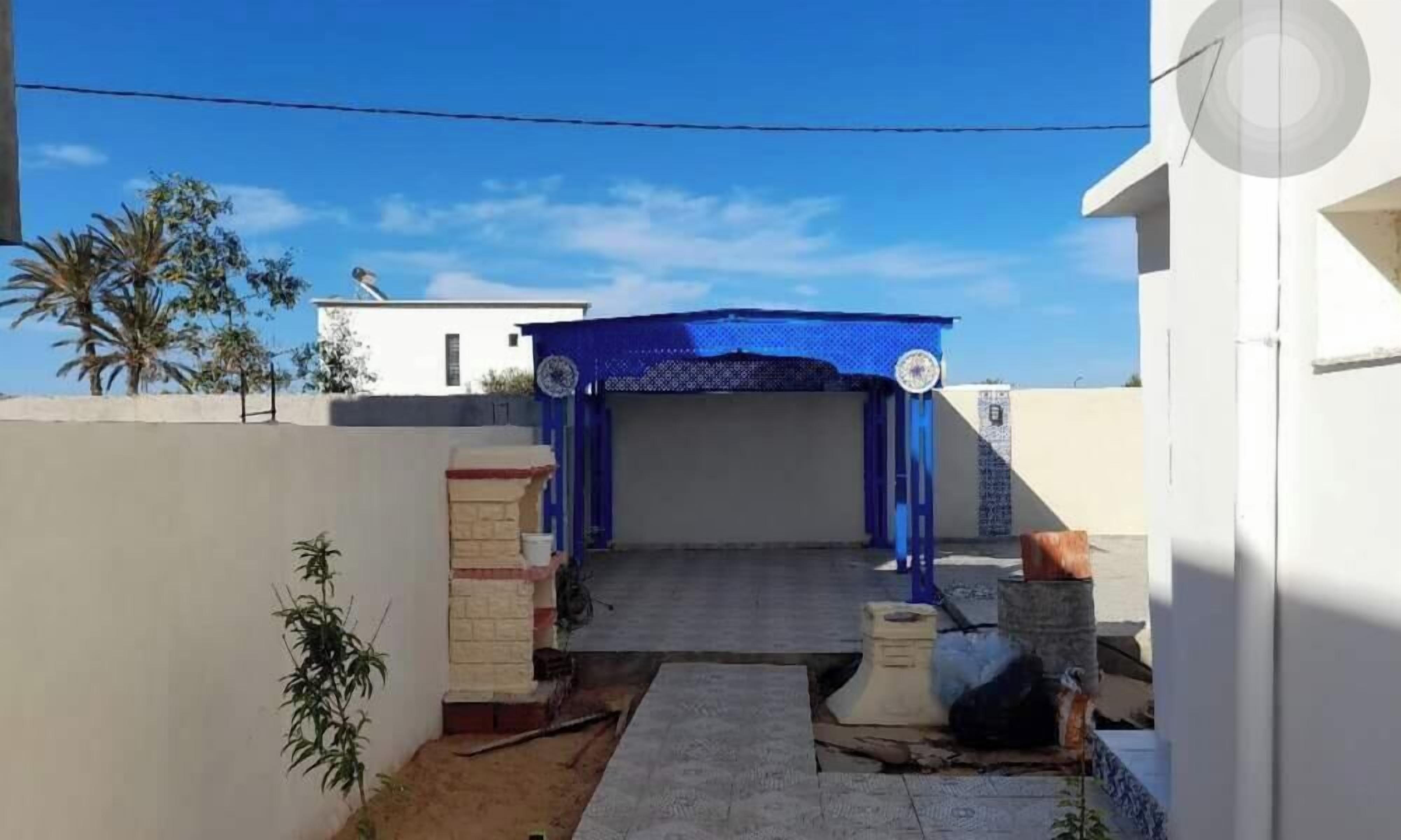 Une entrée fermée par un portail, surmontée d’un auvent bleu, une petite construction en briques et une plante de bambou.