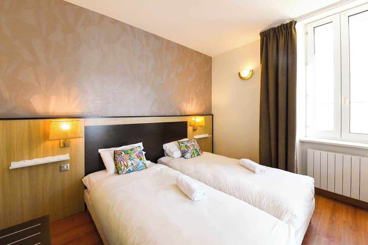 A hotel room with two beds, a wooden headboard, a wall-mounted shelf, and a window with curtains.
