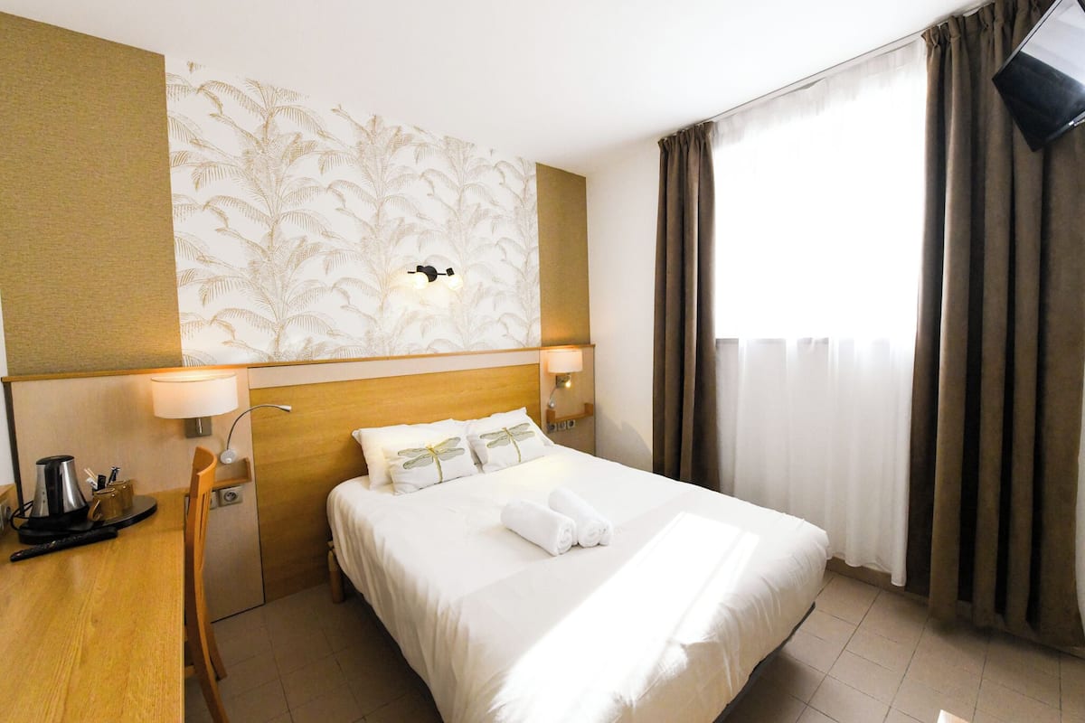 A hotel room with a bed, a desk with a coffee maker, a lamp, and a wall-mounted TV.