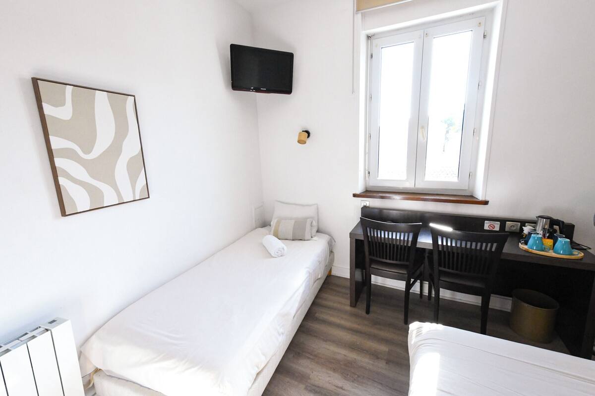 A small, clean room with a single bed, a desk with a chair, a television mounted on the wall, and a window providing natural light.