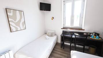 Una habitación pequeña y limpia con una cama individual, un escritorio con silla, un televisor montado en la pared y un ventanal que proporciona luz natural.