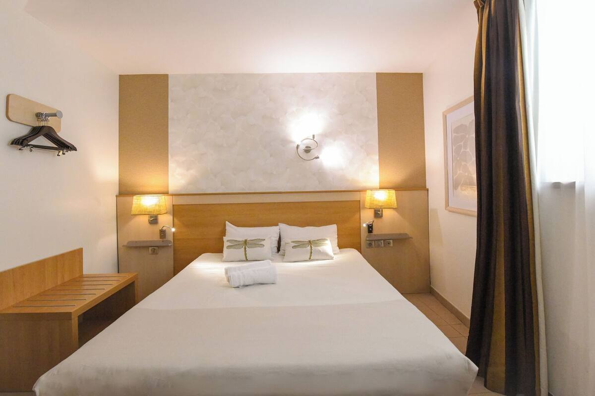 A hotel room with a large bed, two bedside lamps, a wooden bench, a wall-mounted light fixture, and a framed picture on the wall.