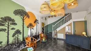 A lobby with a staircase, wicker ceiling lamps, a reception desk, and a mural of trees. -  Hotel le Coureau (Saint-Trojan-les-Bains)
