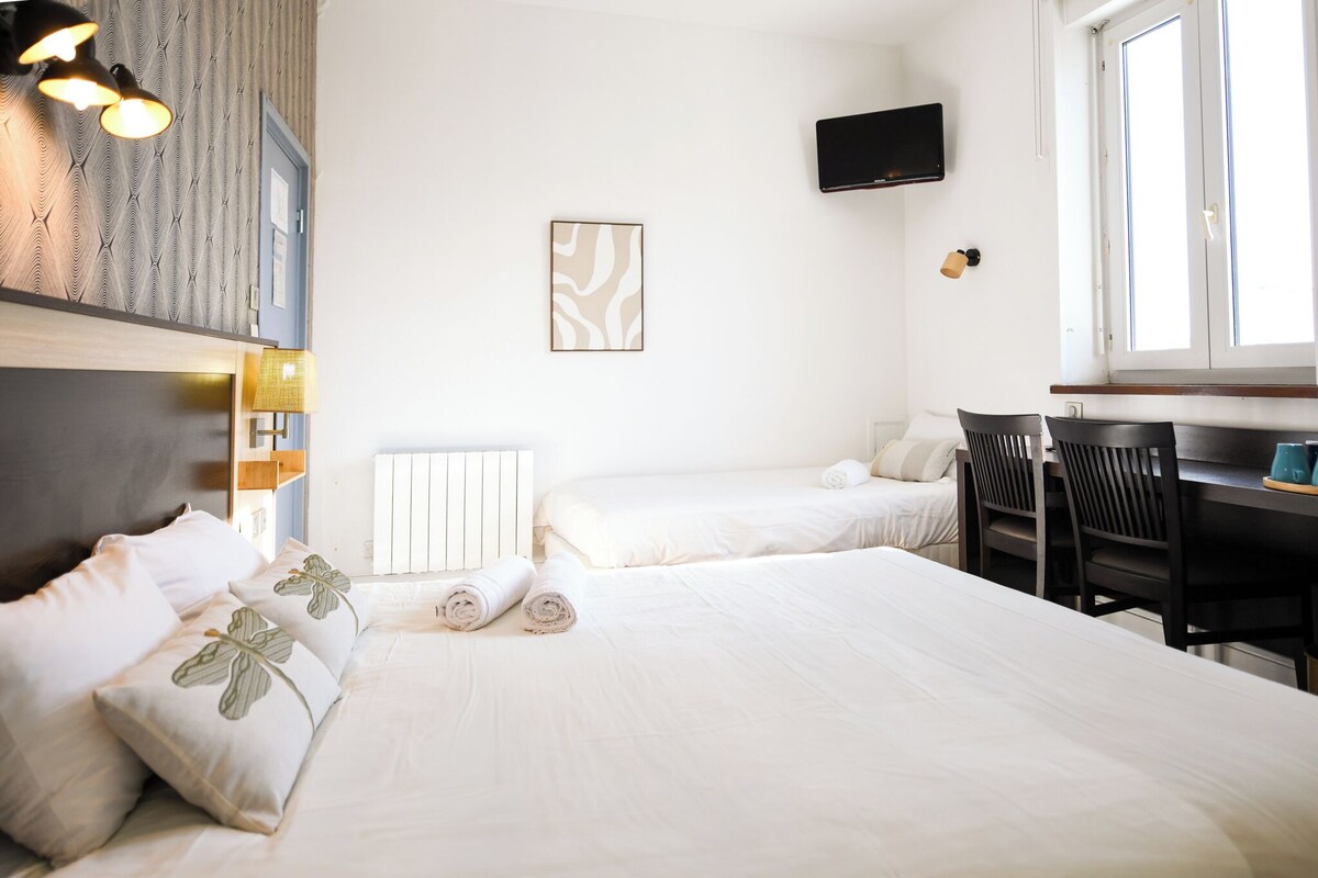 A hotel room with a large bed, two smaller beds, a desk, a chair, a flat-screen TV, and a window with natural light.