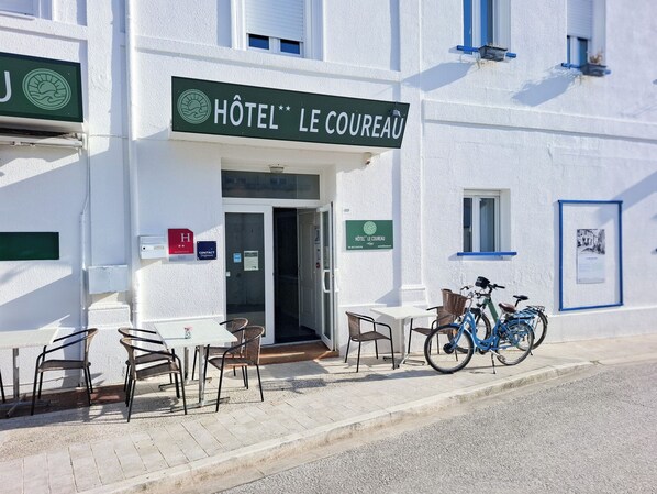 A hotel entrance with a sign reading "Hôtel Le Cœurau," outdoor seating with chairs and tables, and a bicycle parked outside. - Hotel le Coureau (Saint-Trojan-les-Bains)