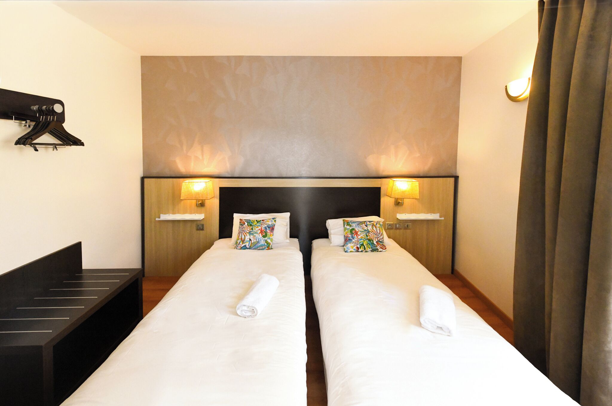 Habitación de hotel con dos camas individuales, cada una con una almohada y una toalla enrollada. Hay lámparas fijadas a la pared a ambos lados de las camas, y un cabecero negro.