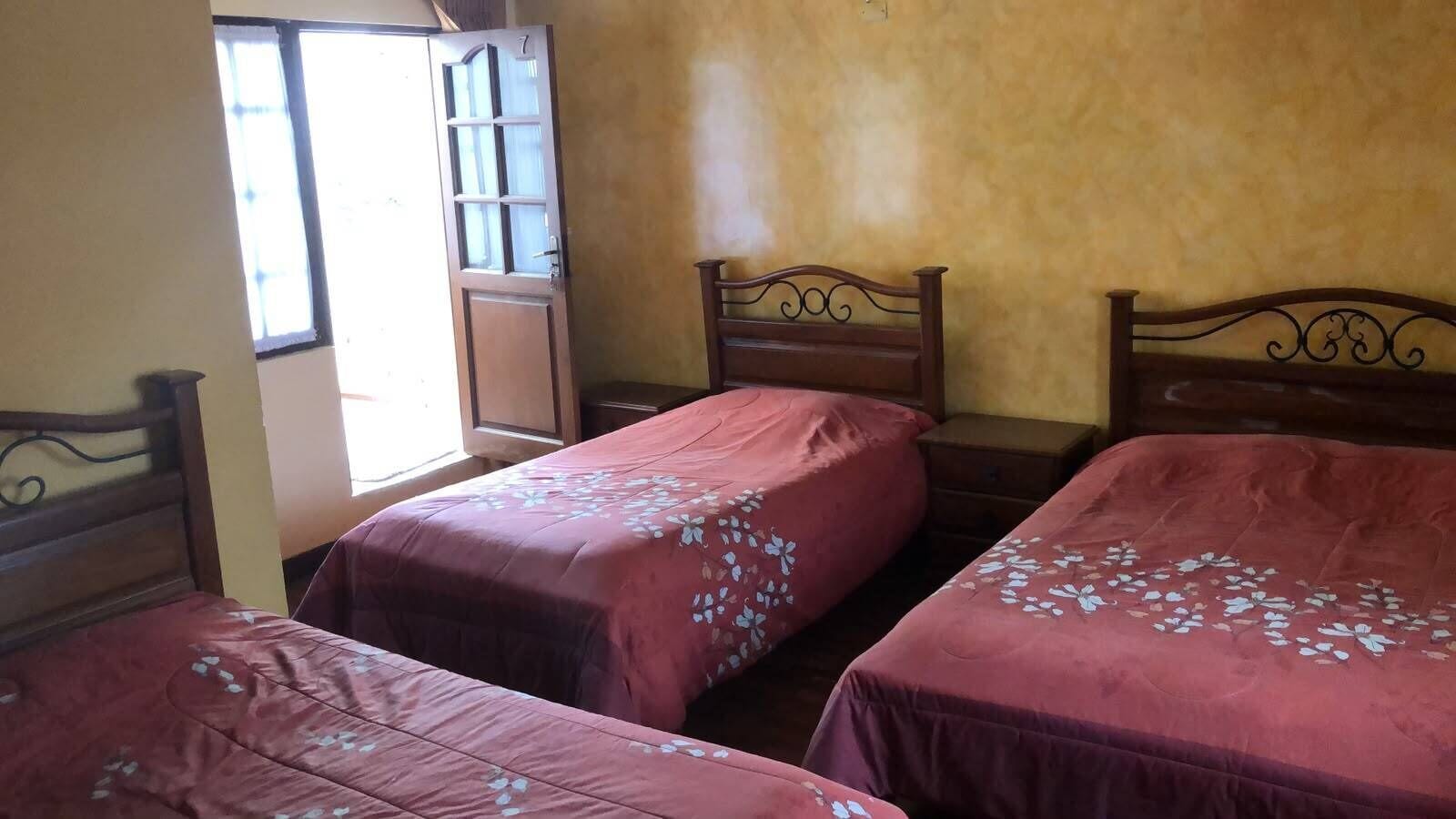 Una habitación con tres camas de madera, cada una con una colcha de motivos florales. Hay una ventana con marco de madera y una puerta con un panel de vidrio.