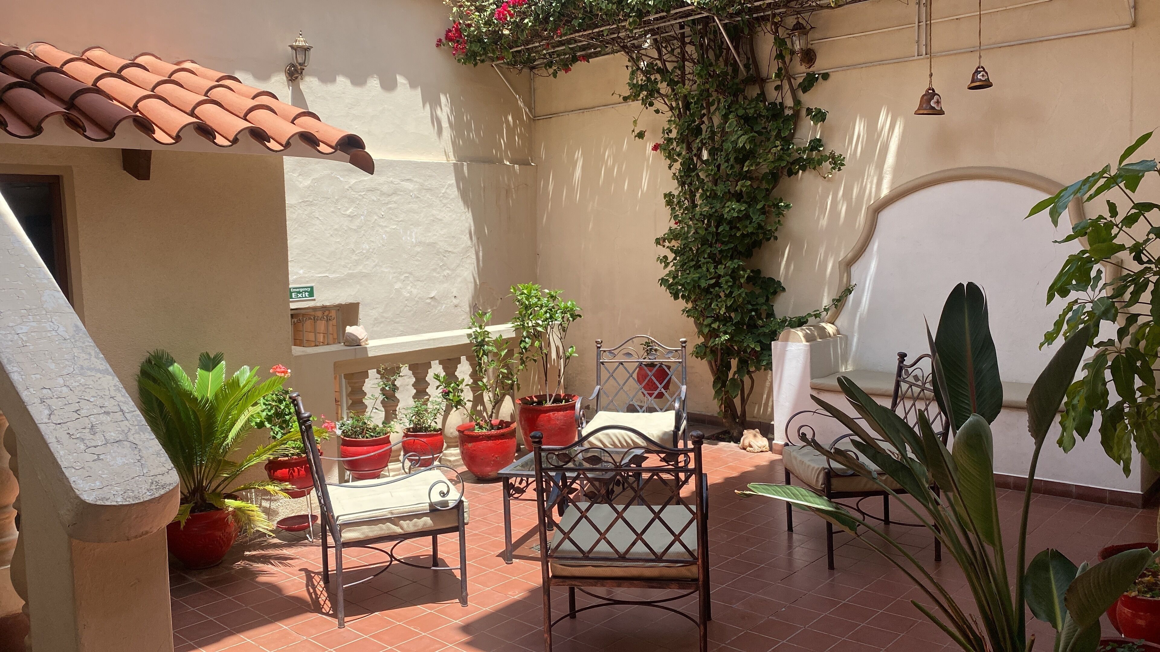 Un patio con suelo de baldosas, un banco de metal, plantas en macetas y un aplique de pared.