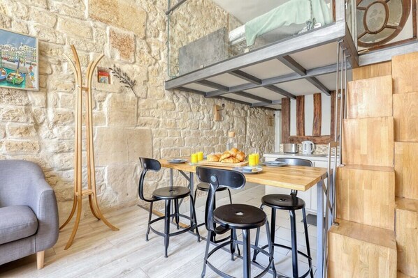 A cozy room with a stone wall, a wooden table with chairs, a sofa, and a kitchen area with a hanging pot rack.