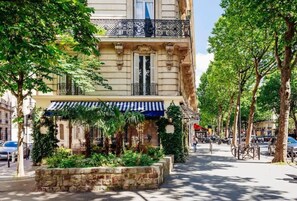 A historic building with a covered outdoor seating area, stone planter, and a street view with trees and parked cars.