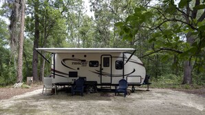 A recreational vehicle (RV) named 'TRACER GIR' parked in a forested area with a table and two chairs in front.