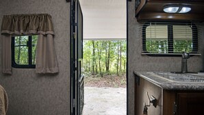 An RV interior with a kitchen area, a window with curtains, and a view of a forested area.